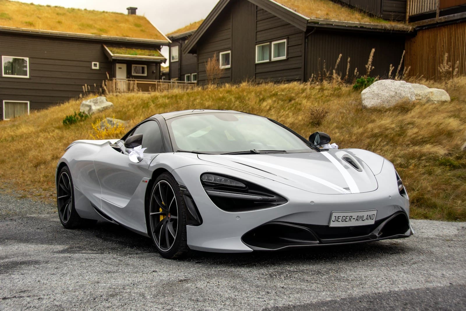 Silver McLaren 720S parked on gravel in front of grass-roof cabins